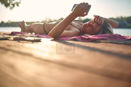 Foto de Atractiva mujer adulta joven sana acostada en un embarcadero de madera por el agua tomando el sol y mirando su teléfono. Viajes, vacaciones, infierno, concepto de estilo de vida. - Imagen libre de derechos