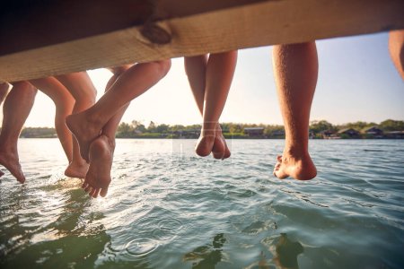 Foto de Amigos junto al lago, sentados en un embarcadero de madera con las piernas colgando al agua, en vacaciones de verano. Viajes, diversión, concepto de estilo de vida. - Imagen libre de derechos