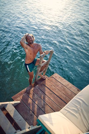 Foto de Joven hombre atlético guapo en traje de baño pantalones cortos que salen del agua, en la escalera a embarcadero de madera. Vacaciones, estilo de vida, concepto salud. - Imagen libre de derechos