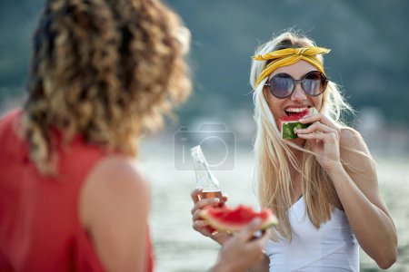 Téléchargez les photos : Deux amies au bord de la mer dégustant un verre et un morceau de melon d'eau. Ensemble, vacances, concept de style de vie. - en image libre de droit