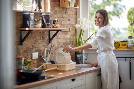 Photo for Joyful young woman washing dishes in a modern kitchen, putting plate away in a dish rack and smiling. Home, lifestyle, family concept. - Royalty Free Image