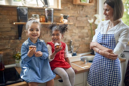 Photo for Two girls, one with fair skin and the other with a radiant dark complexion, sit side by side in the kitchen, savoring delicious muffins.Standing beside them, their mother beams with pride and love. - Royalty Free Image