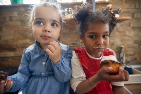 Téléchargez les photos : Gros plan portrait de deux belles petites filles manger des muffins désordonnés ensemble, l'air mignon. Maison, famille, concept de style de vie. - en image libre de droit