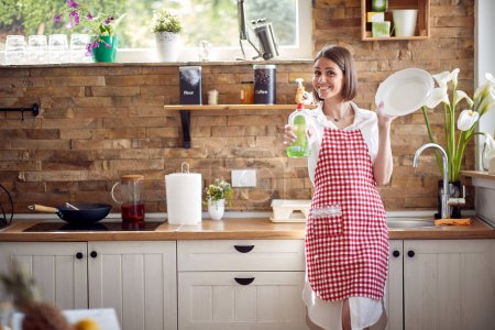 Photo for Attractive young housewife wearing checkered apron standing in the kitchen holding a clean plate and demonstrating favourite dishwashing detergent and smiling. Home, lifestyle concept. - Royalty Free Image