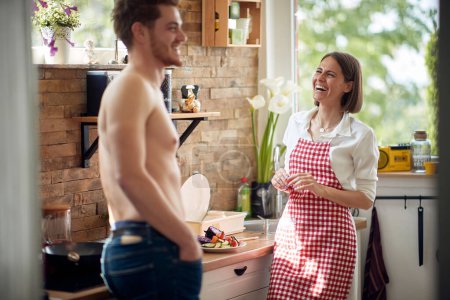 Téléchargez les photos : Jeune femme au foyer debout dans la cuisine avec son mari sexy torse nu parlant et se sentant joyeux ensemble. Maison, famille, concept de style de vie. - en image libre de droit