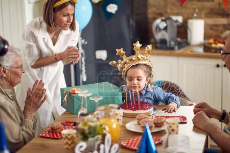 Foto de Linda niña de cumpleaños con la corona inflable soplando velas en su pastel, con su familia a su alrededor, sentada en la mesa de la cocina animándola. Hogar, familia, concepto de celebración. - Imagen libre de derechos