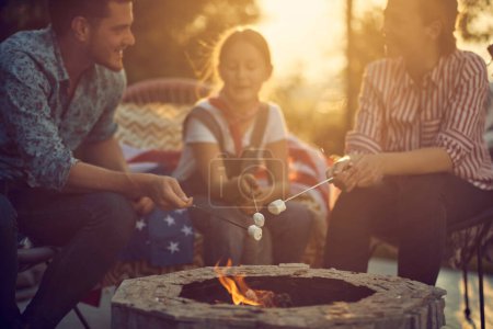 Photo for Beautiful family of three roasting marshmallows together by a fireplace, sitting in cozy chairs outdoors. Togetherness, family, lifestyle concept. - Royalty Free Image