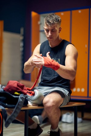 Photo for Handsome young man sitting in dressing room, focusing while tying tape around his hand for protection, preparing for boxing workout. - Royalty Free Image