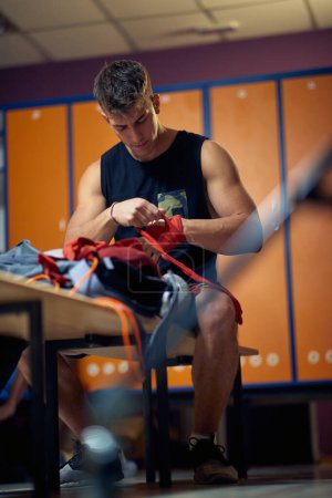 Photo for Young handsome man sitting in gym dressing room tying red tape around his hand. Martial arts, sport, health concept. - Royalty Free Image