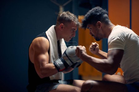 Photo for Handsome young boxing player sitting in dressing room with personal trainer getting ready for workout. Coach encouraging player. Martial arts, sports, lifestyle concept. - Royalty Free Image