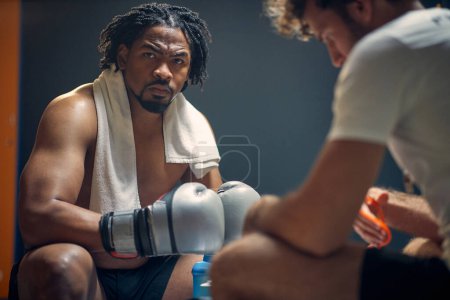 Photo for Powerful strong young boxing player with his coach, getting prepared for workout, sitting on bench face to face. Sports, lifestyle concept. - Royalty Free Image