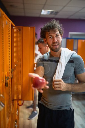 Foto de Joven guapo extendiendo la mano y sosteniendo manzana en el vestuario del gimnasio, bocadillo rápido y saludable después del entrenamiento. Deportes, salud, concepto de estilo de vida. - Imagen libre de derechos