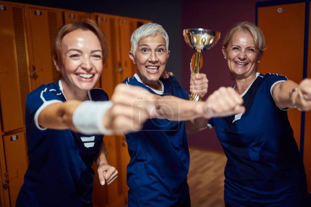 Téléchargez les photos : Équipe sportive féminine de trois personnes célébrant ensemble dans les vestiaires, tenant une coupe dorée. Équipe senior gagnante. - en image libre de droit