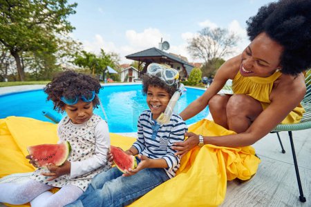 Téléchargez les photos : Famille afro-américaine au bord de la piscine. La mère et ses enfants, un garçon et une fille, s'assoient ensemble sous le soleil chaud. Les enfants se délectent délicieusement de tranches juteuses de pastèque. - en image libre de droit
