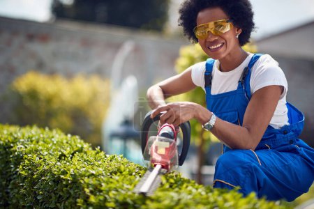 Photo for Afro-American woman as she expertly trims hedges using a Hedge Trimmer. With a confident and focused demeanor, she skillfully shapes the foliage, creating clean and defined lines. - Royalty Free Image