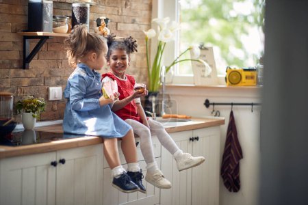 Foto de Dos niñas adorables sentadas en la encimera de la cocina, disfrutando de un aperitivo juntos, comiendo una sandía y un panecillo. Hogar, estilo de vida, concepto familiar. - Imagen libre de derechos