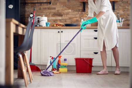 Photo for Faceless shot of young woman wearing gloves, cleaning in the kitchen, moping the floor, with chemical bottles in the background. Home, cleaning, lifestyle concept. - Royalty Free Image