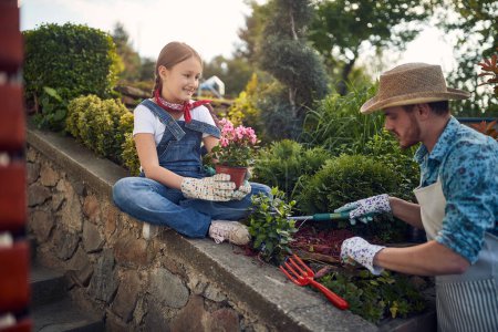 Téléchargez les photos : Père et sa fille unissent leurs forces dans le jardin. La petite fille tient un pot de fleurs avec soin, tandis que son père prépare habilement un trou dans le sol pour la plantation. - en image libre de droit