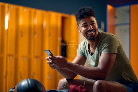 Téléchargez les photos : Jeune homme souriant utilisant un smatphone dans le vestiaire de la salle de gym avant l'entraînement. Sport, actif, concept lifestyle. - en image libre de droit