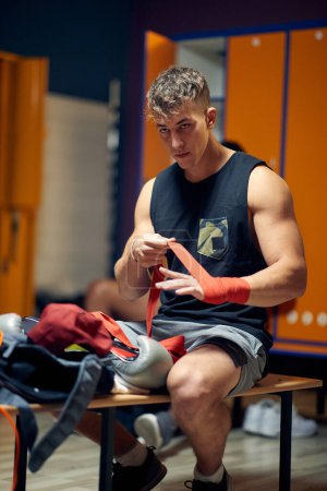 Photo for Young man in dressing room, sitting on bench, tying red tape around his hand, getting ready for workout. Boxing, martial arts, sports concept. - Royalty Free Image