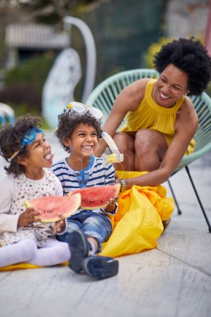 Photo for Loving mother is captured alongside her children, a girl and a boy, enjoying a delightful moment by the pool. The summer sun illuminates their smiles as they indulge in juicy watermelon slices. - Royalty Free Image