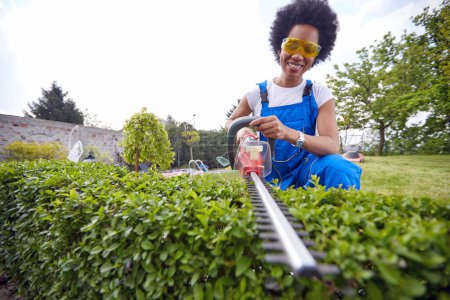 Photo for Woman confidently wields a precision trimmer as she skillfully shapes a vibrant living hedge. With a steady hand and focused gaze, she effortlessly navigates through the lush foliage. - Royalty Free Image