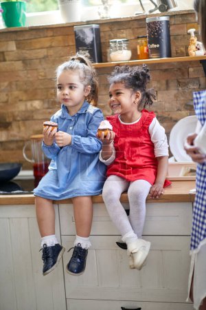 Photo for Two young girls are captured in a delightful moment as they sit on the kitchen counter, savoring delicious cupcake muffins. With smiles of pure delight and anticipation, they hold the cupcakes. - Royalty Free Image