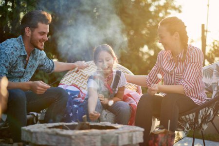 Photo for Family gathers in their backyard, basking in the joy of togetherness. The parents and their daughter are seen indulging in the simple pleasure of roasting marshmallows over a crackling fire. - Royalty Free Image