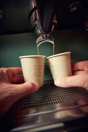 Foto de Manos masculinas caucásicas sosteniendo dos tazas de papel debajo del chorro de café negro - Imagen libre de derechos