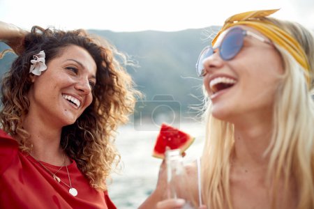 Foto de El primer plano de dos amigas jóvenes está disfrutando de una jugosa sandía y una bebida en el muelle en un hermoso día soleado en la playa. Amistad, vacaciones, mar - Imagen libre de derechos