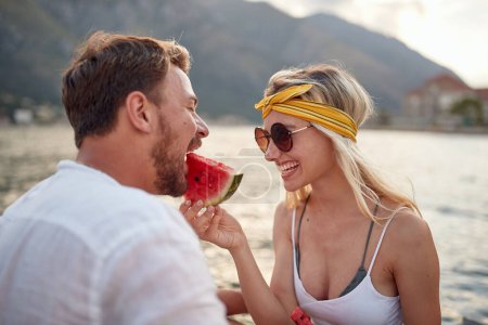 Photo for Close-up of a young couple feeding each other with a juicy watermelon while they sitting on the dock on the seaside on a beautiful day. Love, relationship, holiday, sea - Royalty Free Image