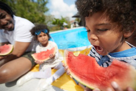 Photo for Young Afro-American boy takes a bite into a slice of watermelon,his face lighting up with delight. In the background, his father and sister hold their own slices, ready to savor the refreshing fruit. - Royalty Free Image