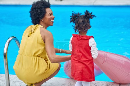 Téléchargez les photos : Joie rayonnante partagée entre une mère afro-américaine et sa fille au bord de la piscine. Tous deux vêtus de robes vibrantes, leurs sourires illuminent la scène alors qu'ils se prélassent dans la chaleur du soleil. - en image libre de droit