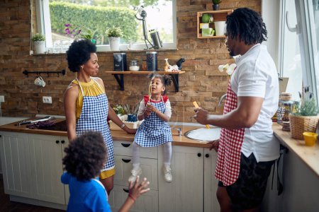 Foto de La familia afroamericana se reúne alrededor del mostrador de la cocina. La madre, el padre, el hijo y la hija están reunidos, con la niña sentada en el mostrador, soplando con entusiasmo burbujas de jabón. - Imagen libre de derechos