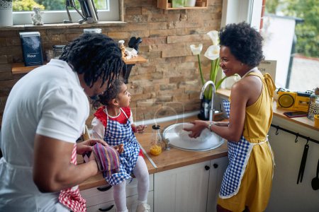 Photo for Family harmony, the mother is carefully washing the dishes in the kitchen while the father attentively dries them. Their little girl sits on the kitchen counter, delighting in a sweet treat. - Royalty Free Image