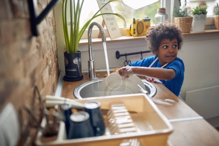 Photo for Young Afro-American boy takes on the responsibility of washing dishes with great enthusiasm. Donning a colorful apron, he stands confidently at the sink, surrounded by a sea of soapy suds. - Royalty Free Image
