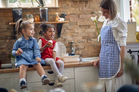 Photo for Two toddler girls sit side by side in the kitchen, delighting in delicious muffins. Their radiant smiles reflect the pure happiness while their mother stands beside them. - Royalty Free Image