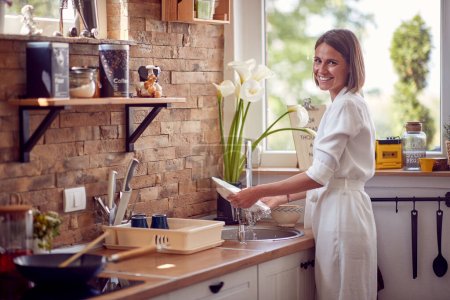 Foto de Foto horizontal de una joven lavando platos en una cocina moderna, sintiéndose alegre. Estilo de vida, concepto casero. - Imagen libre de derechos