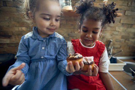 Foto de Primer plano retrato de dos niñas desordenado comer magdalenas en la cocina juntos, sintiéndose feliz. Hogar, familia, concepto de estilo de vida. - Imagen libre de derechos