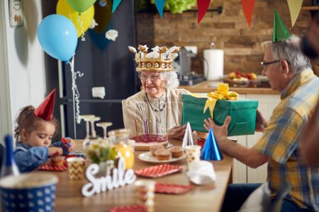 Photo for Joyful senior woman recieving a birthday gift from husband, sitting at kitchen table with their granddaughter, baloons and ornaments in backgorund. Family, lifestyle, senior life concept. - Royalty Free Image