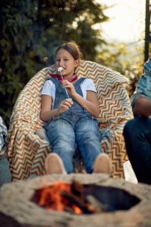 Foto de Hermosa niña comiendo malvaviscos a la parrilla, sentada al aire libre junto a una chimenea en una silla acogedora, disfrutando del día de verano en el patio. Concepto de estilo de vida. - Imagen libre de derechos