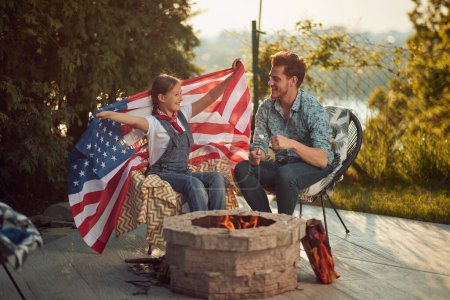 Photo for Cheerful girl and father sitting outdoors by a fireplace in their yard roasting marshmallows, holding USA flag. Independece day, fourth of July concept. - Royalty Free Image