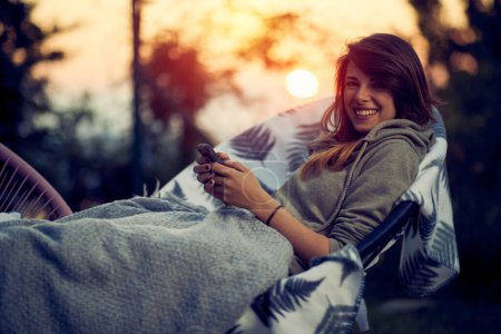 Foto de Joven mujer alegre relajándose en una silla acogedora con una manta, sosteniendo un teléfono y sonriendo. Fondo de puesta de sol. - Imagen libre de derechos