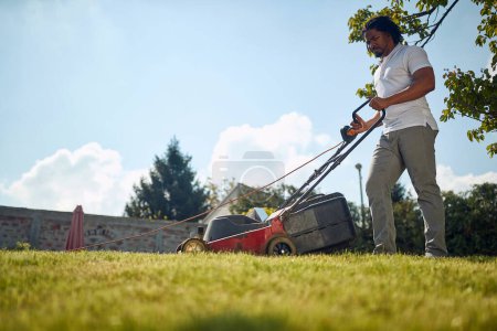 Photo for Handsome young african american man mowing grass outdoors, making the backyard beutiful and neat. Lifestyle, activity, home concept. - Royalty Free Image