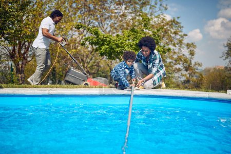 Foto de Madre y su hijo pueden ser vistos limpiando diligentemente la piscina con una red de skimmer, trabajando juntos como un equipo.En el fondo, el padre se puede ver cortando el césped. - Imagen libre de derechos