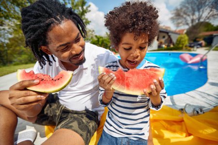 Foto de Padre e hijo afroamericanos sentados junto a la piscina, disfrutando de una jugosa sandía. Con risa y deleite, su energía alegre es cautivadora. - Imagen libre de derechos