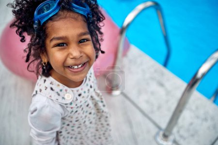 Photo for Afro-American girl by the poolside. With a vibrant smile illuminating her face, she exudes excitement and happiness. She has dive goggles perched on her head. - Royalty Free Image