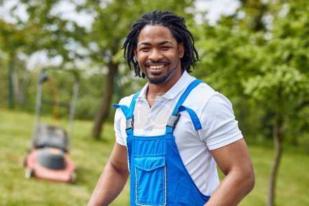 Téléchargez les photos : Un jardinier afro-américain en gros plan, portant un uniforme et un sourire rayonnant. Debout sur un fond d'herbe verte luxuriante et tondeuse à gazon. - en image libre de droit