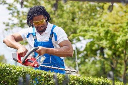 Foto de Joven hombre afroamericano en traje de trabajo con trimmer eléctrico para cortar setos al aire libre, jardinería. Hogar, estilo de vida, concepto de jardinería. - Imagen libre de derechos