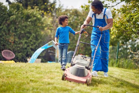 Photo for Father and son outdoors in the backyard mowing grass with grass mower, son helping his dad in gardening. African american family. - Royalty Free Image